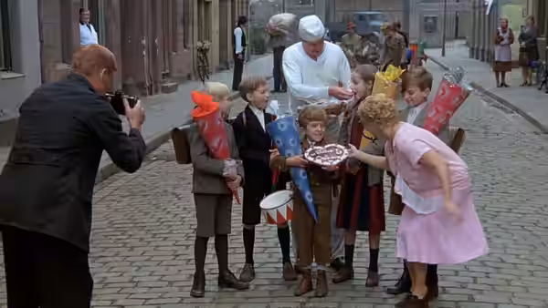 A photographer captures schoolchildren gathered around a cake on a cobbled street