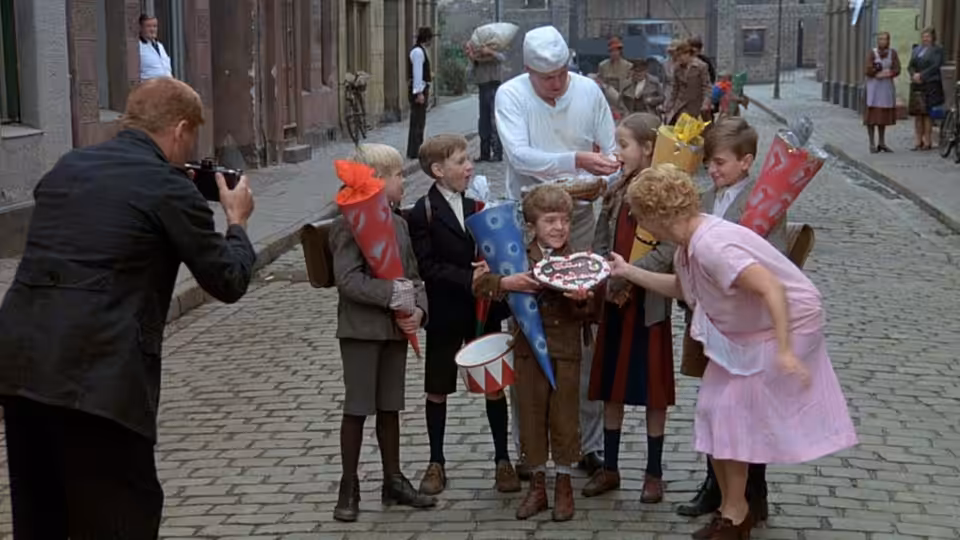 A photographer captures schoolchildren gathered around a cake on a cobbled street