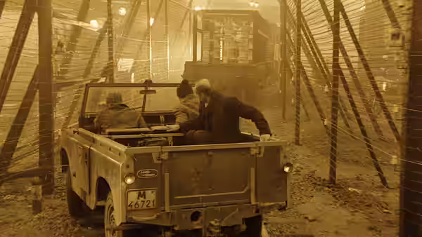 Three men drive a jeep along a fenced road in an industrial zone