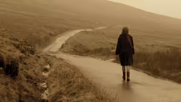 Bess (Emily Watson) walking along a wet road through open hilly countryside