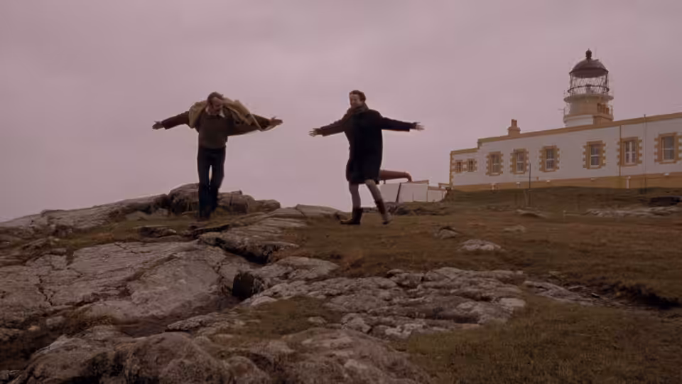 Jan (Stellan Skarsgård) and Bess (Emily Watson) with arms outstretched on a rocky hillside near a lighthouse
