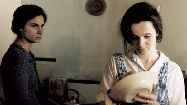 Bess (Emily Watson) holding a bowl as Dodo (Katrin Cartlidge) stands nearby indoors