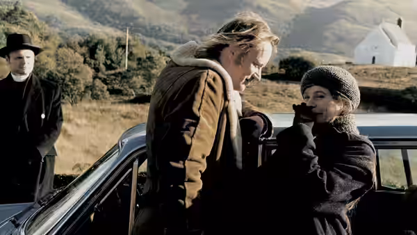 Jan (Stellan Skarsgård) leaning toward Bess (Emily Watson) by a car while a man watches