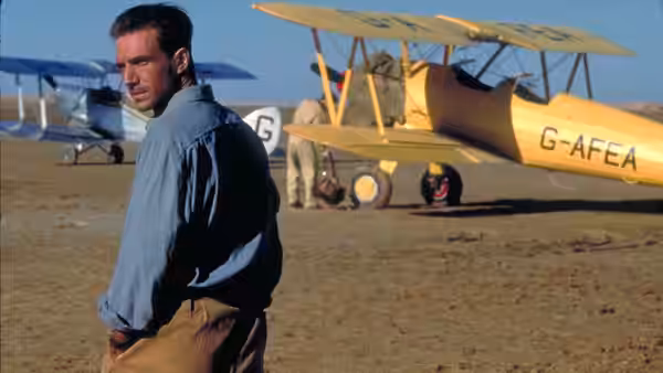 Almásy (Ralph Fiennes) stands with hands in pockets beside a yellow biplane