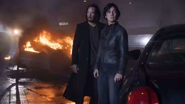 Neo (Keanu Reeves) and Trinity (Carrie-Anne Moss) stand beside a car as wreckage burns behind them
