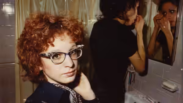 A red-haired woman in glasses poses as another woman looks in a bathroom mirror