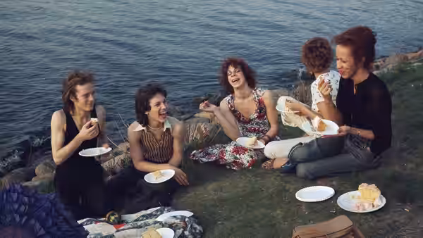 Five women share food on a grassy bank beside the water