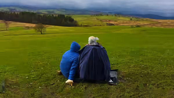 A blue-hooded figure and a gray-haired figure sit together, looking across green fields