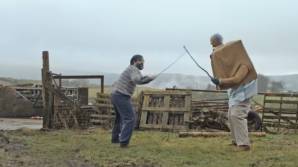 Brian (David Earl) and Charles (Chris Hayward) spar with sticks in a farmyard