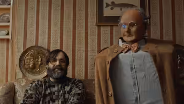 Brian (David Earl) sits beside Charles (Chris Hayward) in a patterned living room