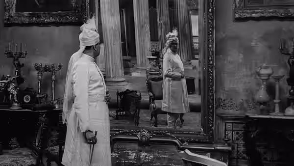 A turbaned man stands in an ornate music room, facing his reflection in a large mirror