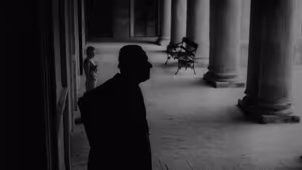 A man stands in silhouette beside a colonnade, with a chair across the courtyard