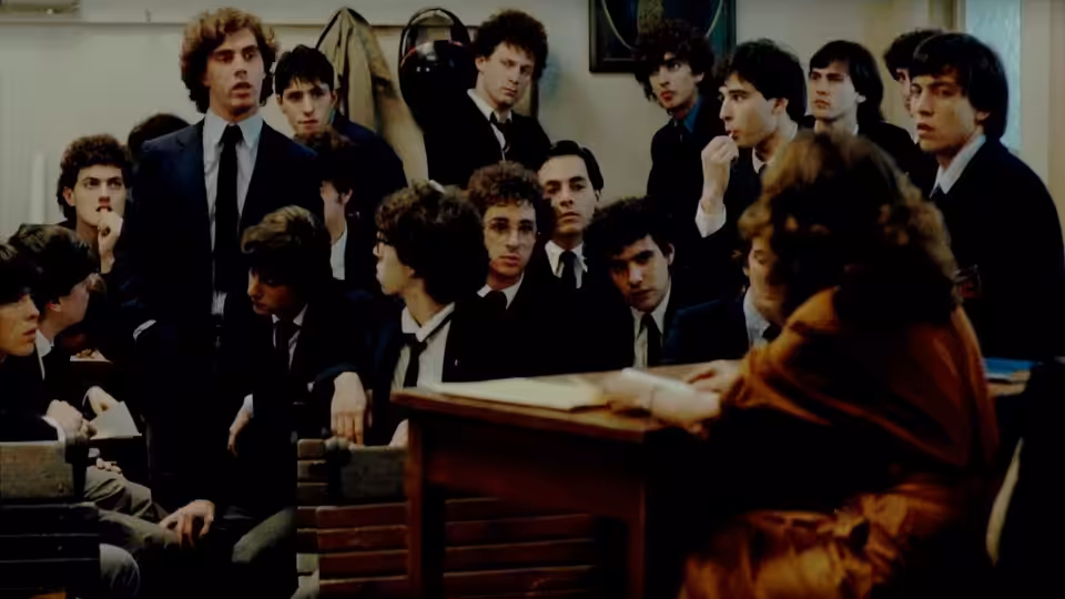 A woman addresses a crowded classroom of male students from behind a lectern