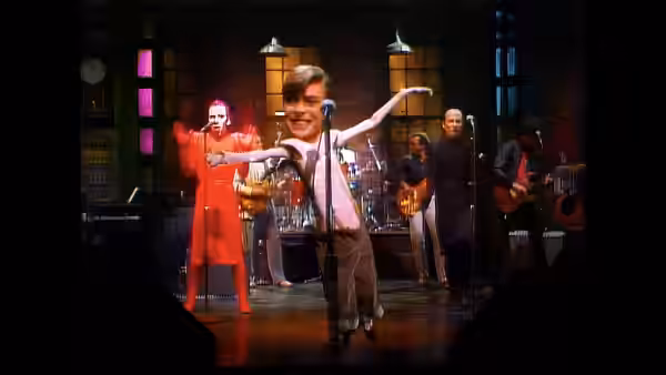 A performer dances at a microphone as musicians play behind him onstage