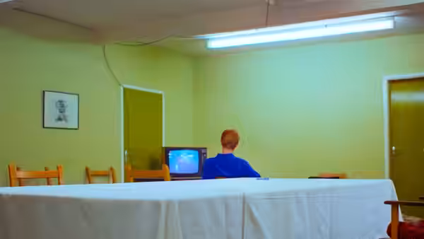 A red-haired man watches a television in a sparsely furnished room