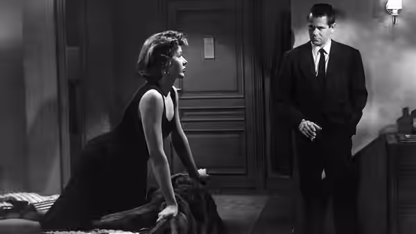 Dave Bannion stands beside a woman seated on a bed in a bedroom