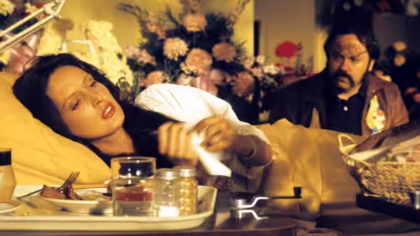 A woman in a hospital bed reaches across her meal tray as a bearded man watches