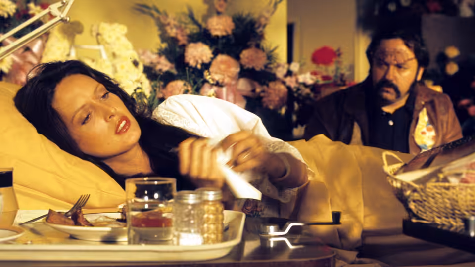 A woman in a hospital bed reaches across her meal tray as a bearded man watches