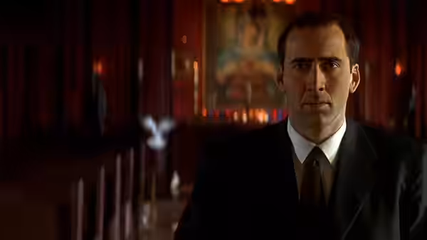 Nicolas Cage in a suit standing inside a dimly lit church with red curtains