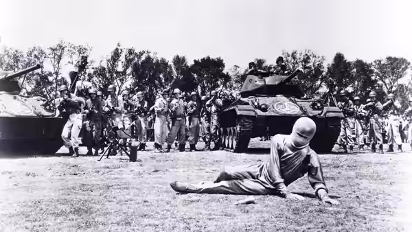 A figure in a silver suit lies on the ground before tanks and a crowd