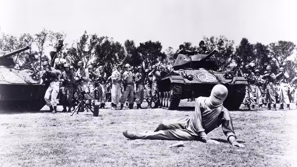 A figure in a silver suit lies on the ground before tanks and a crowd