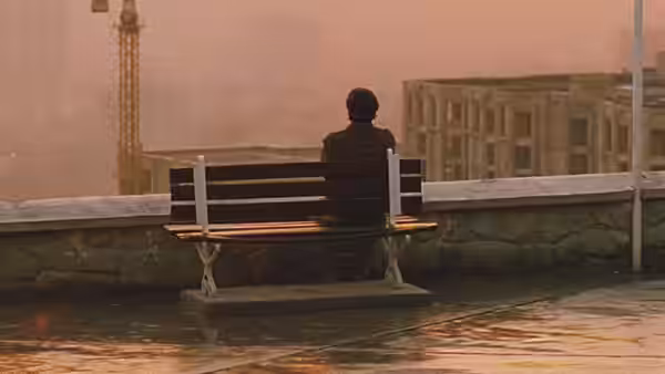 Man in dark jacket seated on bench overlooking hazy city with crane