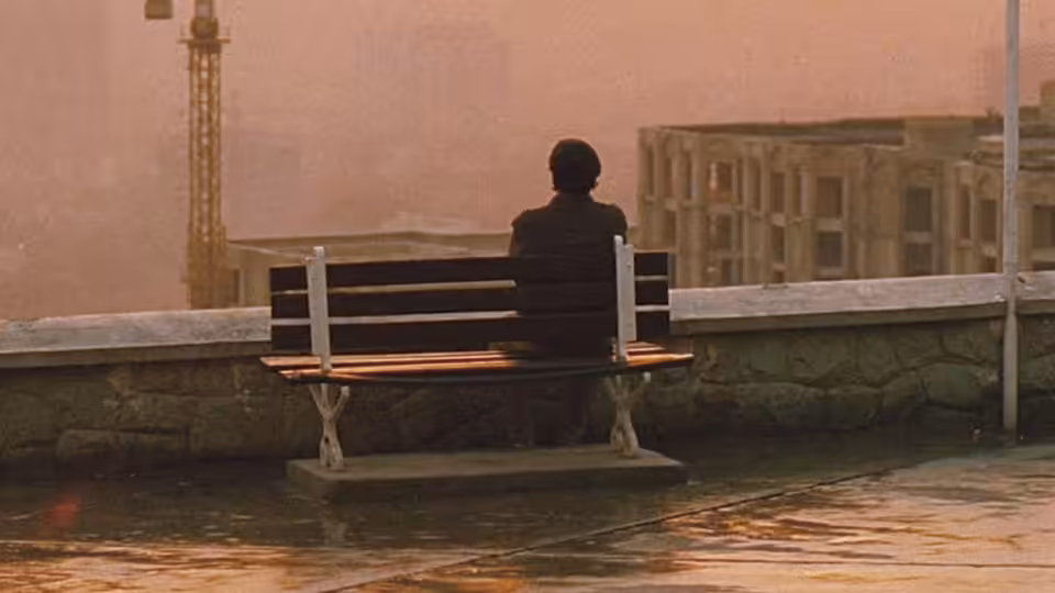Man in dark jacket seated on bench overlooking hazy city with crane