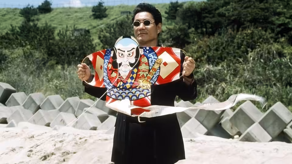 Yoshitaka Nishi (Takeshi Kitano) holding a large kite on the shore