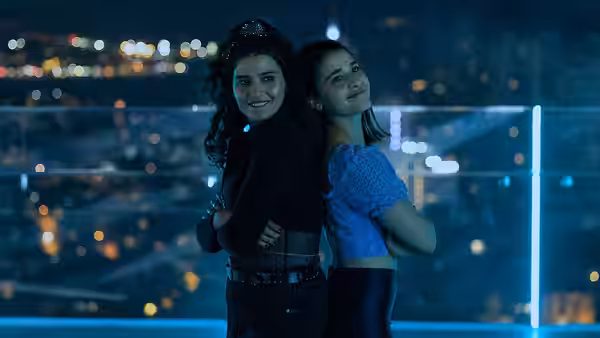 Two young women stand back to back on a balcony overlooking city lights