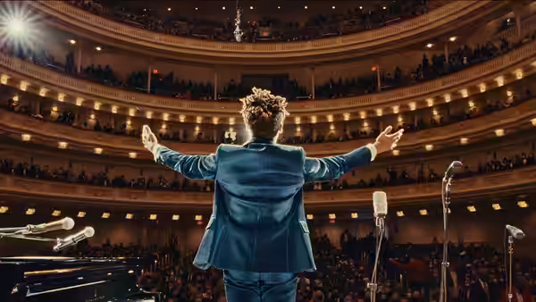 Jon Batiste stands with arms outstretched before a packed concert hall audience