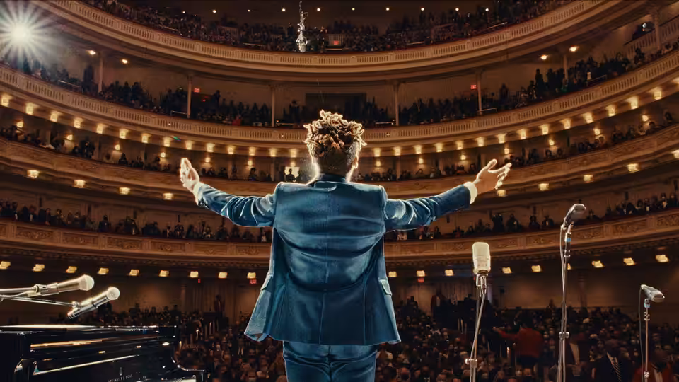 Jon Batiste stands with arms outstretched before a packed concert hall audience