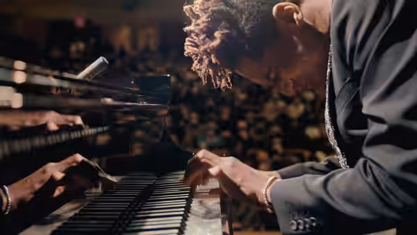 Jon Batiste leans over a piano, playing keys onstage before an audience