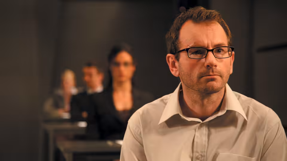 White (Luke Mably) watches tensely from his desk as other candidates sit behind him