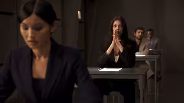 Chinese Girl (Gemma Chan) sits in the foreground as a woman clasps her hands at a desk