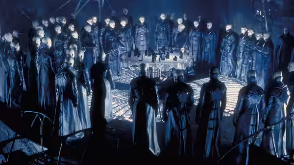 Pale bald figures in dark coats standing around an illuminated circular table