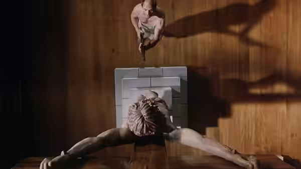 A man lies spread-eagled on a wooden floor as another crouches nearby