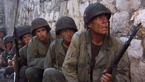 The Sergeant (Lee Marvin) crouches with fellow soldiers beside a stone wall