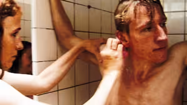 Two young men in a tiled bathroom one shirtless and touching his ear