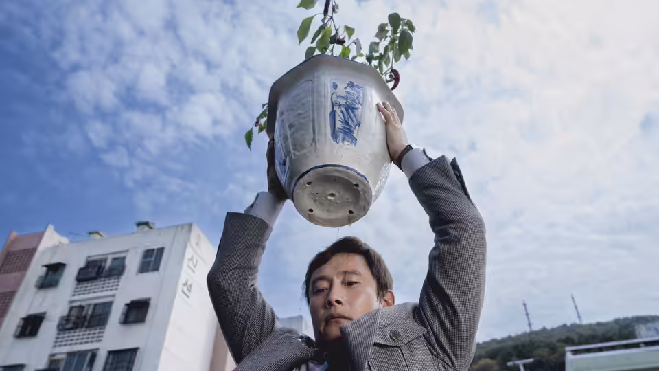Man-su (Lee Byung-hun) lifting a large potted plant overhead outdoors