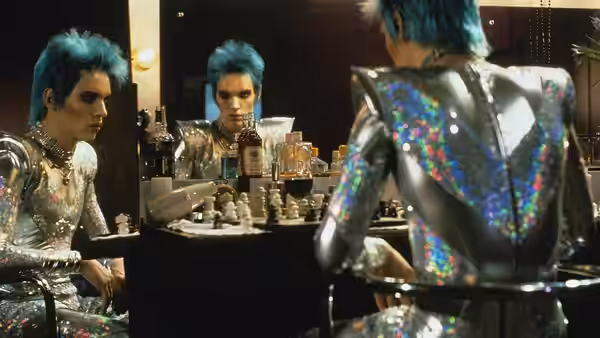 Man with blue hair in sequined costume sits at vanity looking in mirror
