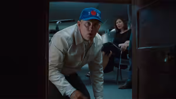 A man in a cap crouching in a doorway with a woman seated behind