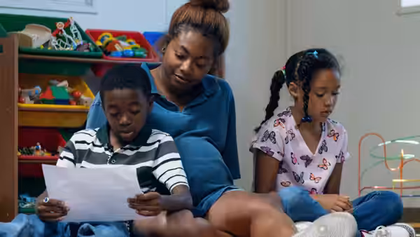 Gia (Tia Nomore) sits with two children as a boy reads a sheet of paper