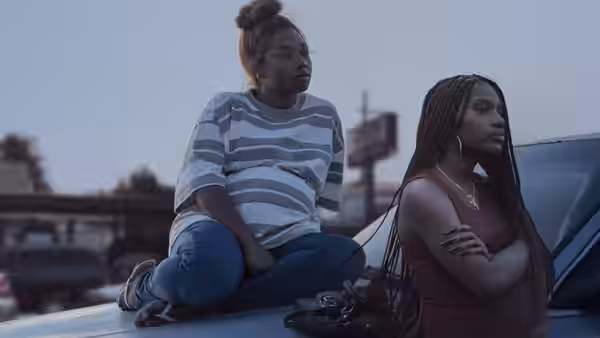 A pregnant woman sits on a car beside a braided woman standing with folded arms