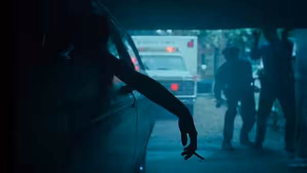 An arm holding a cigarette extends from a car near an ambulance at night
