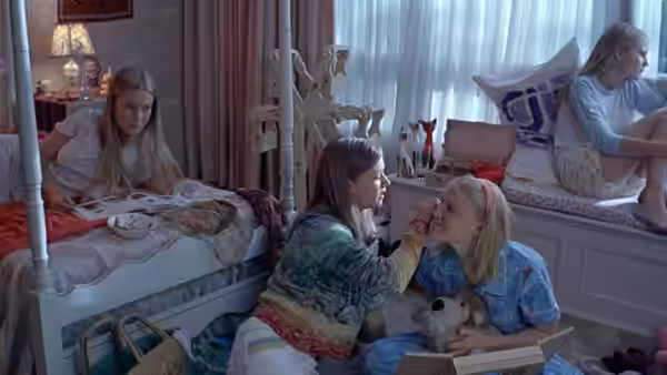 Girls sit in a bedroom while one applies makeup to another's face