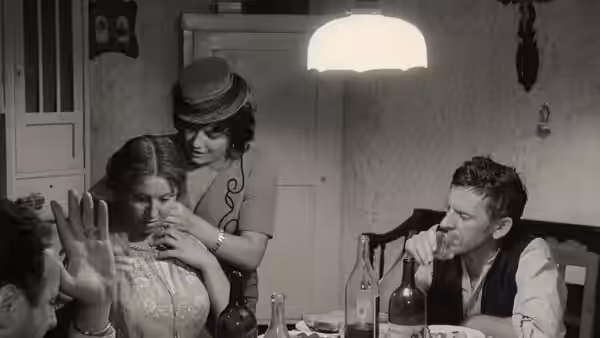 A man drinks at a table while a woman in a hat supports another woman