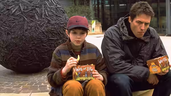 Marcus Brewer (Nicholas Hoult) and Will Freeman (Hugh Grant) eat snacks beside a sculpture
