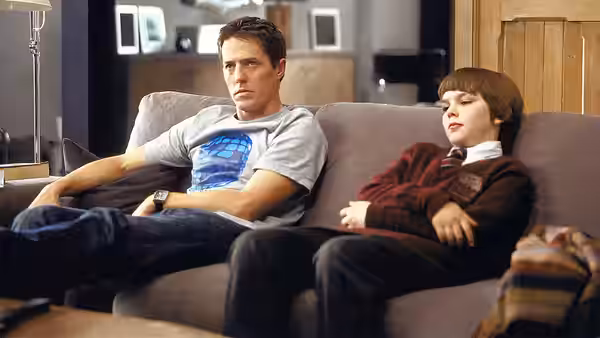 Will Freeman (Hugh Grant) and Marcus Brewer (Nicholas Hoult) sit side by side on a sofa