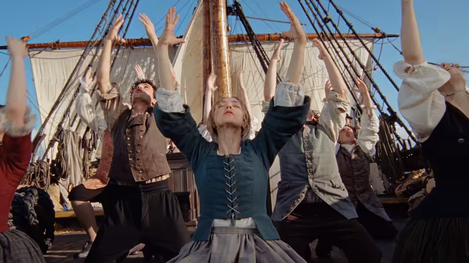 Ann Lee (Amanda Seyfried) raising her arms with others on a ship's deck