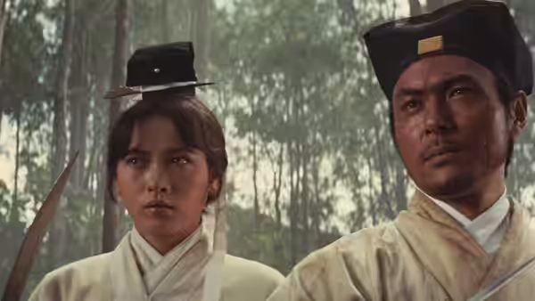 A woman and a man in pale robes stand together in the forest